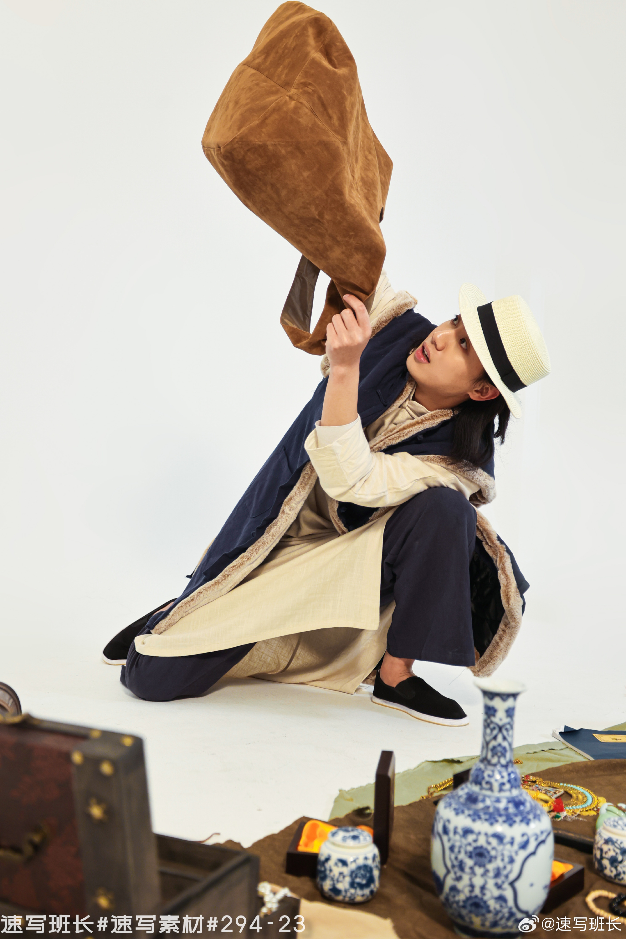 a man wearing a long, traditional asianstyle coat is dramatically swinging a large, brown shoulder bag. he is crouched low to the ground and looking intently at something out of the frame. the scene is set in a studio with a white background, and several props are scattered around the man, including a chest, a vase, and books.
