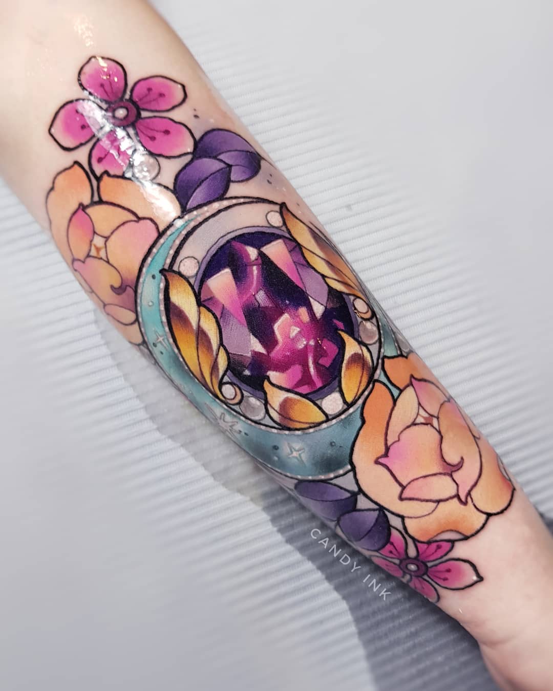 a neo traditional tattoo of a purple crystal on someone's forearm. the crystal is surrounded by a gold crescent moon decorated with pearls. there are pink and yellow flowers around the moon. the tattoo artist's signature "candy ink" is written below the tattoo in small grey letters.