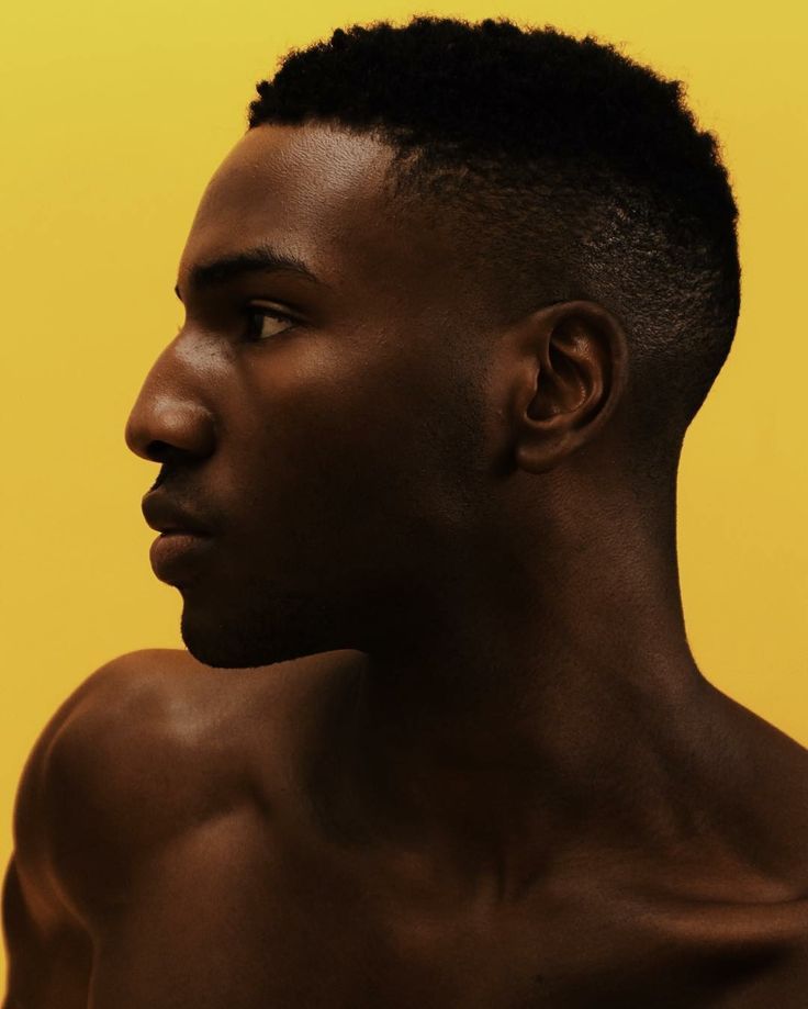 a portrait of a darkskinned man in profile against a yellow background. he has a short fade haircut and bare shoulders.