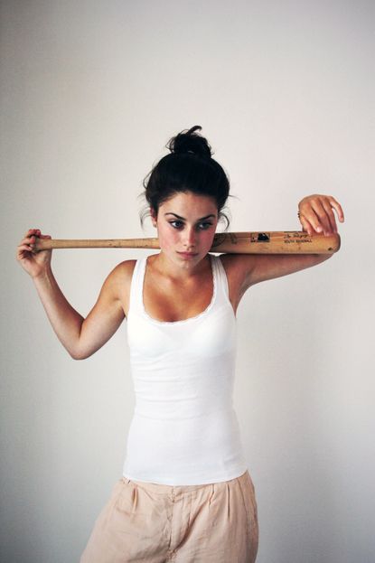 a young woman with brown hair pulled back in a bun, is wearing a white tank top and beige pants and is holding a wooden baseball bat over her shoulder. she's looking directly at the camera with a serious expression on her face.