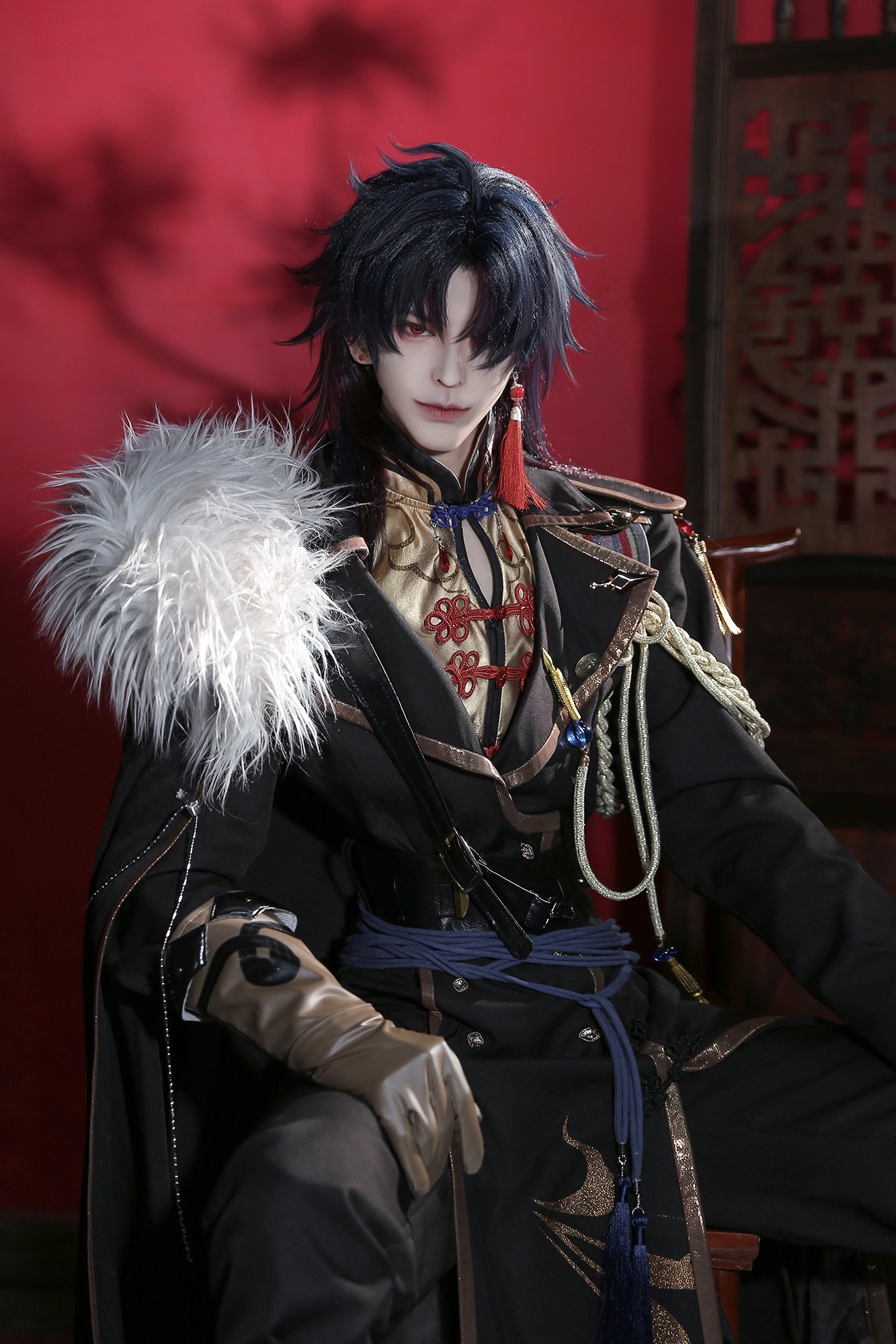 a young man in a dark and elegant cosplay, with long black hair and a white fur stole over his shoulder, sits in a traditional chinese setting. his costume has ornate gold embellishments, a red and gold sash, and a black sword strapped to his side. his expression is serious and his pose is confident.