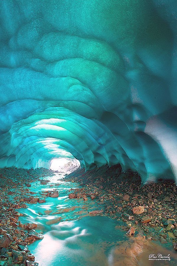 a stunning image of an ice cave with a stream flowing through it. the cave is illuminated with a bright light at the end, creating a tunnellike appearance. the ice is a mesmerizing shade of blue, and the stream adds a sense of movement and life to the scene. the rocky ground and stone debris surrounding the stream highlight the natural and rugged beauty of the location.