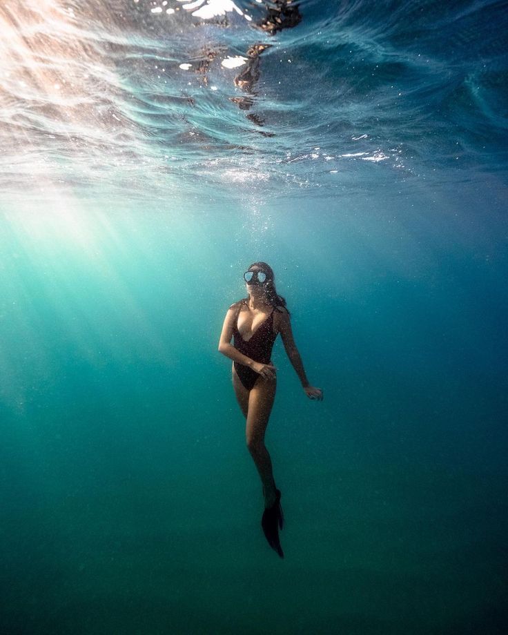 an underwater view of a woman in a black swimsuit, swimming with a dive mask and fin. the woman is swimming towards the surface of the ocean, where sunlight is shining.  the image is captured from a low angle perspective.