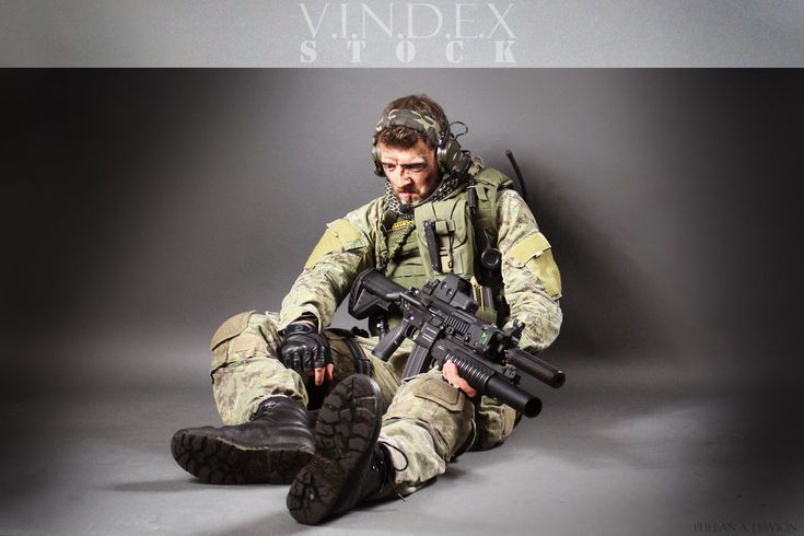 a soldier in full military camouflage sits on the ground with a rifle, and a serious expression on his face.  he has a tactical vest,  a headset on and a lot of combat gear.  the image is dark, which lends itself to the seriousness of the military profession.