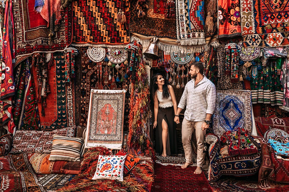 a man and woman are holding hands in front of a shop filled with rugs. the man is lightskinned and has a beard and is wearing a striped shirt and khakis. the woman is fairskinned and has long, dark hair. she is wearing a white top and a long black skirt with a slit. they are both barefoot. the shop is filled with colorful rugs in a variety of patterns. there are also cushions and other textiles in the shop.