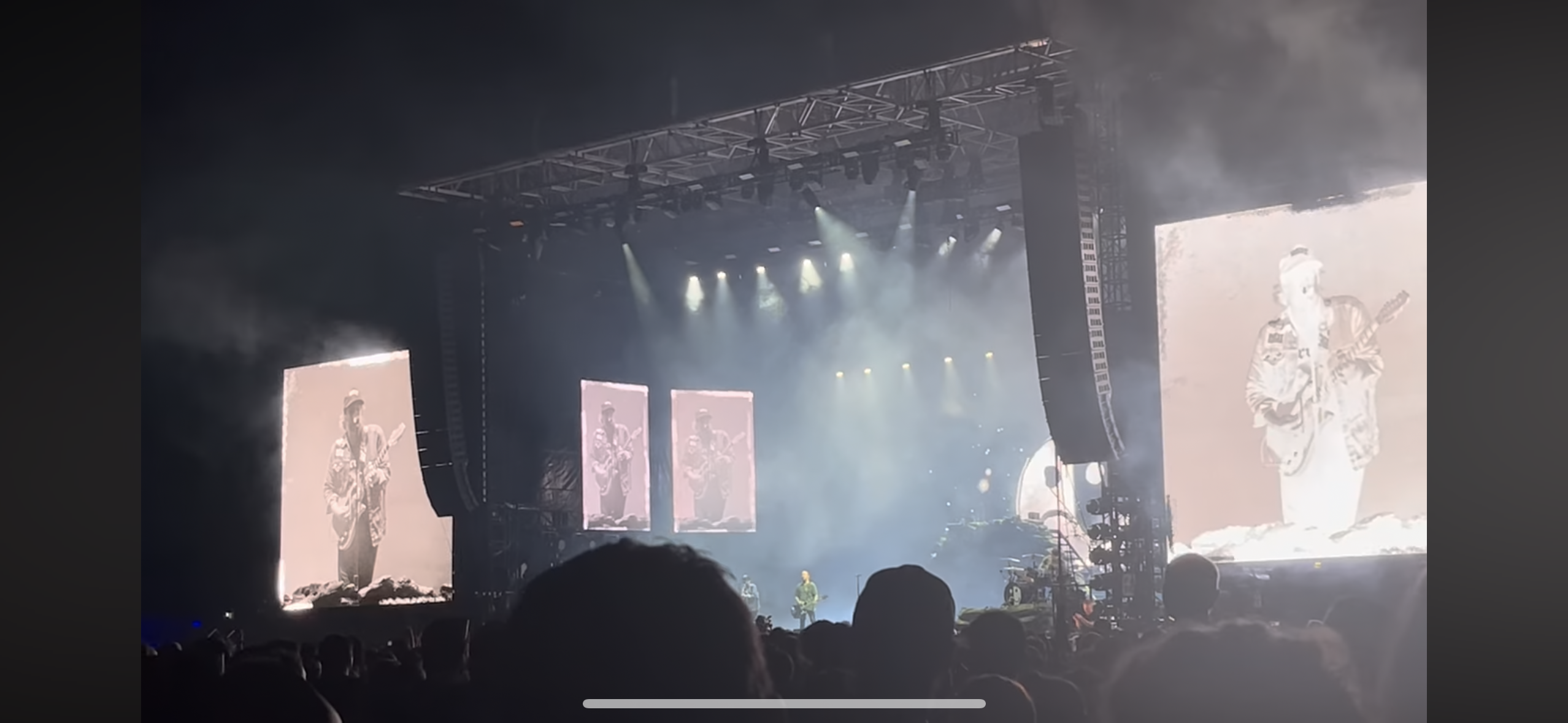 this image shows a concert stage with a band performing on stage. there is a large crowd of people in the audience watching the show. the stage is lit with bright lights and there are large screens playing videos of the band. there is also a singer performing on stage, with a guitar in hand. the image is taken from the point of view of someone in the audience.