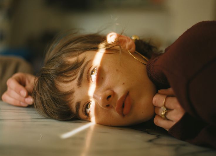 a young woman lies on her side with her head resting on her hand, she is looking toward the camera with a slightly serious expression on her face.  she has long brown hair and bangs, and her face is lit by a soft, golden light.  she is wearing a maroon sweater and has a golden ring on her finger.
