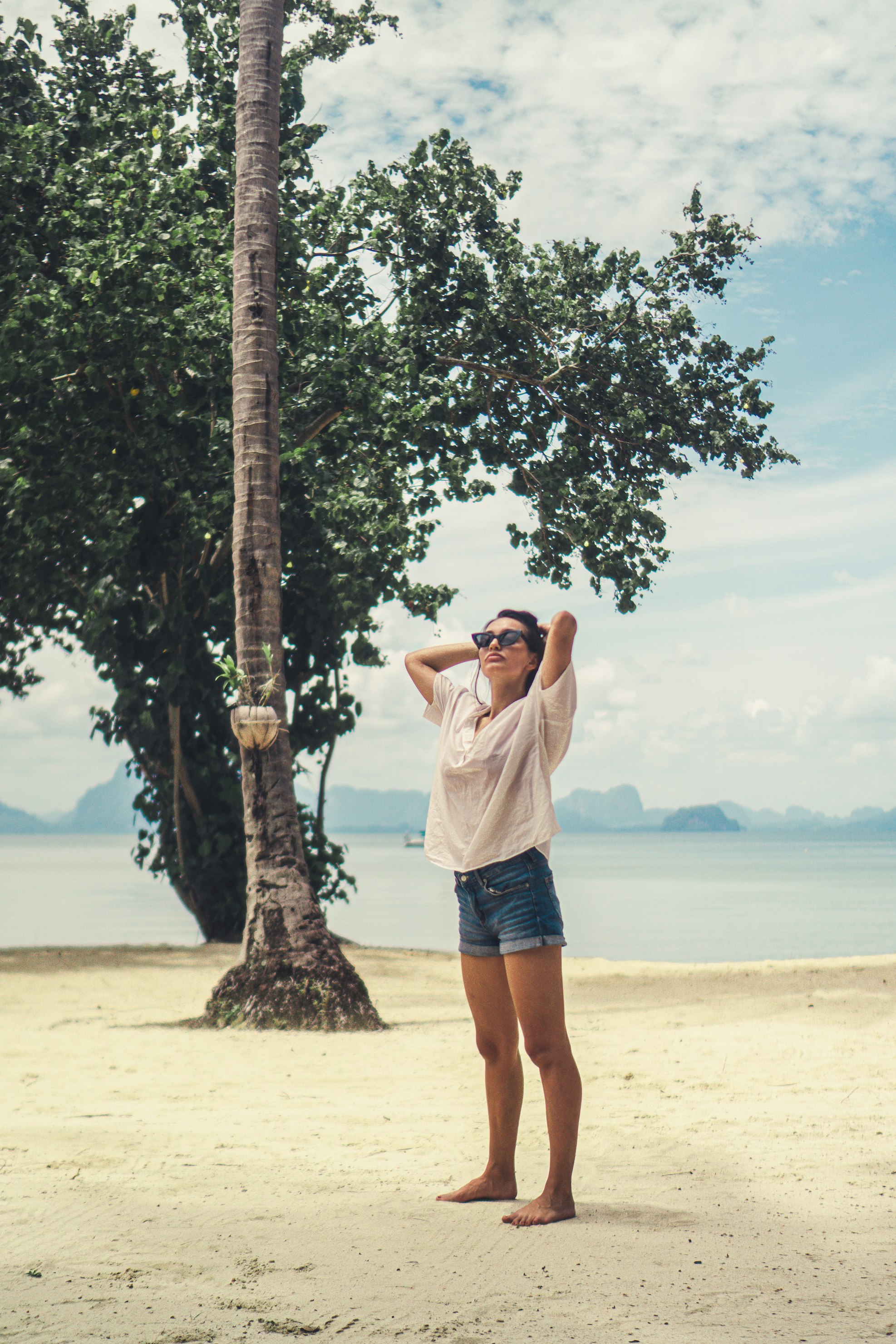 a young woman in denim shorts and a white shirt stands on a sandy beach with her arms raised above her head, she's wearing sunglasses. behind her are lush green tropical trees and a calm ocean, indicating a tropical destination. the overall atmosphere is relaxed and serene. the setting suggests a summer vacation or holiday.