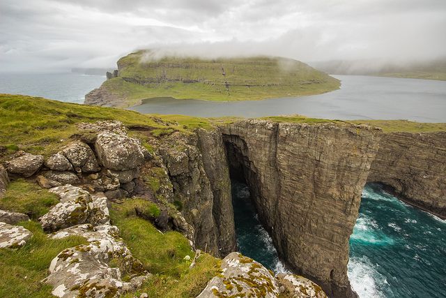 a dramatic view of a sea cliff on the faroe islands, with a lake in the background and a sea stack jutting out in front of it. the cliff is sheer and high, with a deep chasm that runs down to the ocean below. the water is a deep blue, and the sky is a hazy gray. the entire scene is bathed in a soft, ethereal light, making it look like something out of a dream. the image is a testament to the power and beauty of nature.