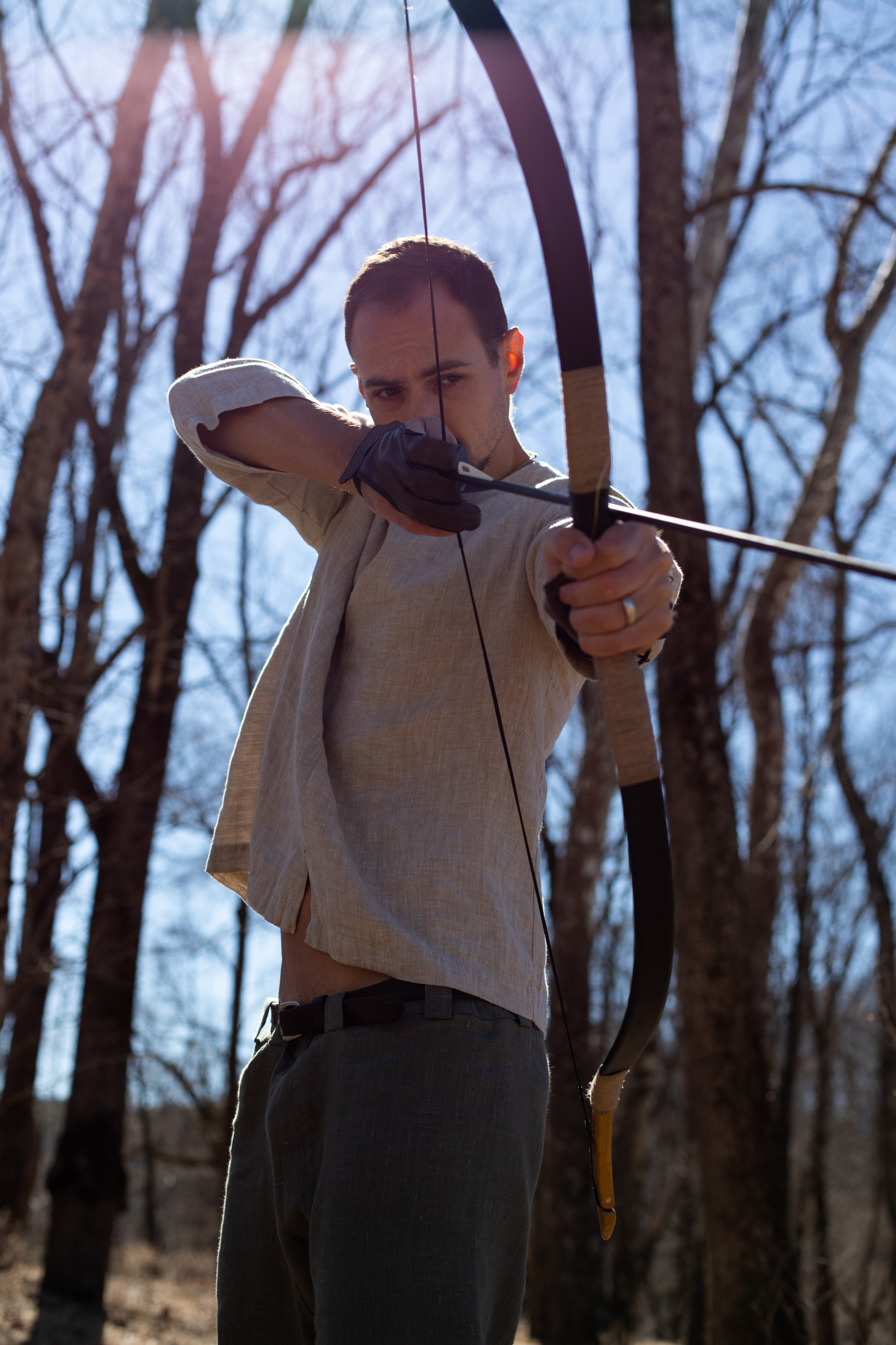 a man stands in a forest, aiming a bow and arrow directly at the viewer. he is wearing a lightcolored shirt and dark pants, and is lit from behind by the sun, creating a silhouette effect. the low angle perspective emphasizes the height and reach of the trees surrounding him.