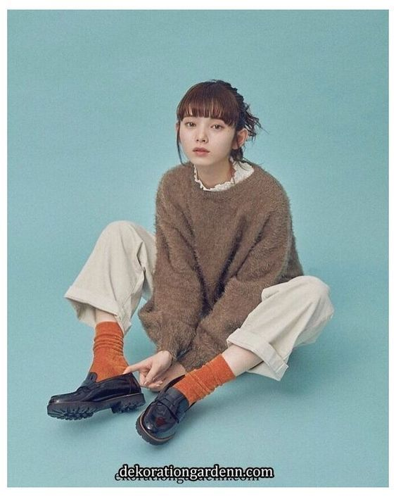 young woman sits on a light blue backdrop, her knees drawn up. she wears a brown fuzzy sweater, white pants, orange socks, and black loafers. she has short brown hair with bangs. her pose is relaxed and natural, with one hand resting on her ankle and the other on her knee. the lighting is soft and diffused, creating a gentle and approachable atmosphere. her expression is calm and pleasant.