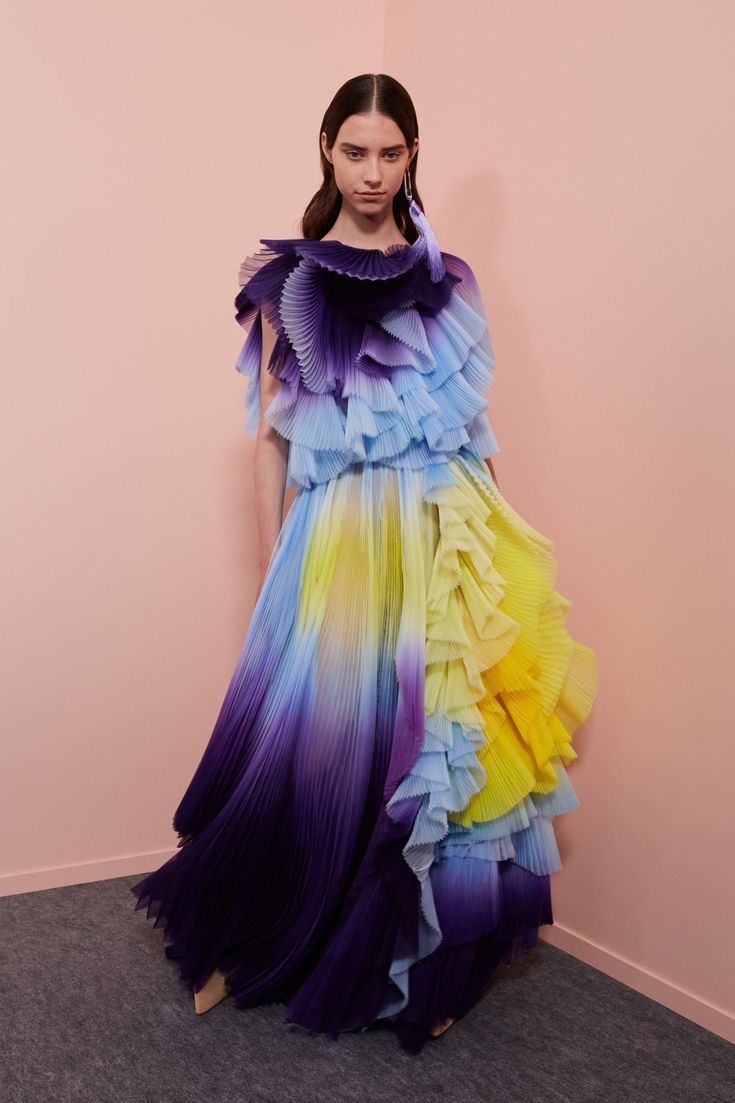 a woman in a purple, blue and yellow ombre pleated dress with ruffles stands in a room with pink walls.