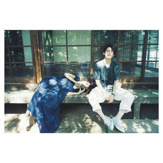 a young couple sits on a wooden bench outside of a traditional japanese house. the man is wearing a white shirt and pants, and the woman is wearing a blue dress. both of them are holding cameras. the photo is taken in natural sunlight, with a vintage feel.