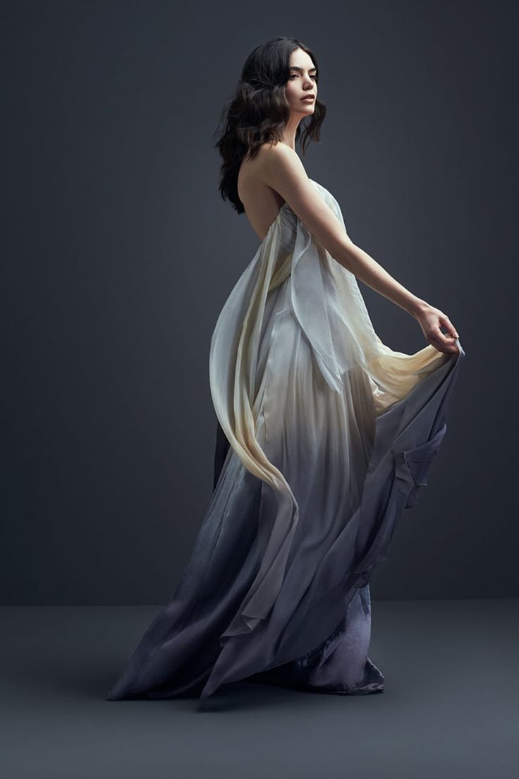 a woman in a beautiful flowy gown stands with her back to the camera and looks over her shoulder. the dress is ombre, starting light and flowing into a dark shade of blue. the woman is in a studio and the background is dark. the fabric of the dress is moving dramatically, almost like it's being blown by wind.