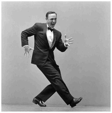 a blackandwhite photo of a man in a tuxedo, dancing against a gray backdrop. the lighting is bright and even, suggesting a studio environment. the man's pose is energetic and expressive, capturing the joy of movement.