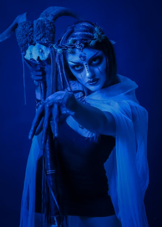 a captivating portrait of a woman in elaborate fantasy costume and makeup. she appears as a dark fantasy elf or sorceress, with pale skin, dark eyes, and face paint that adds to her mystical appearance. she is holding a staff with horns, adding to the otherworldly feel. the blue lighting enhances the dramatic and mysterious mood. the woman's intense gaze and pose add to her powerful and enigmatic presence.