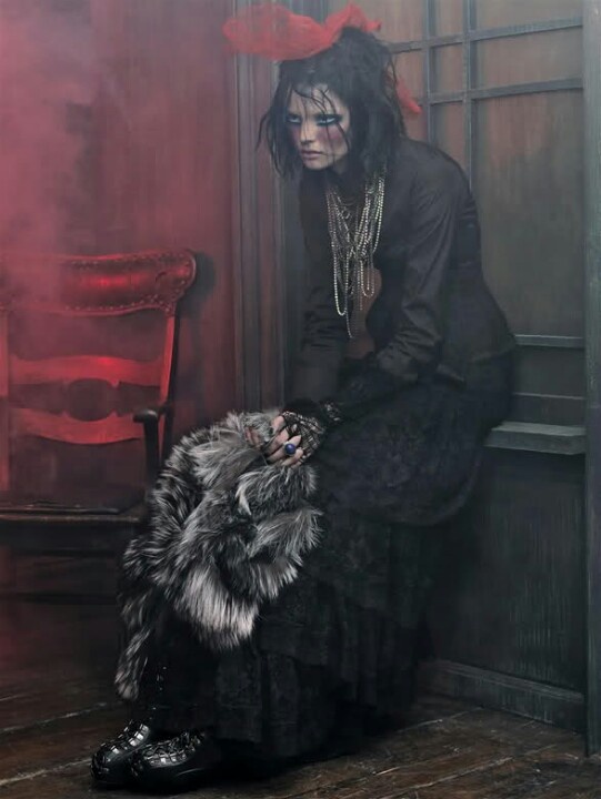 a woman with dark hair and dramatic makeup sits in a dimly lit room. she is dressed in a gothic style, wearing a black dress, multiple necklaces, and holding a fur stole. she has a hat with a large red bow. behind her is an old chair and a window, with a red smoke effect adding to the atmosphere. the overall mood is gloomy and vintage.