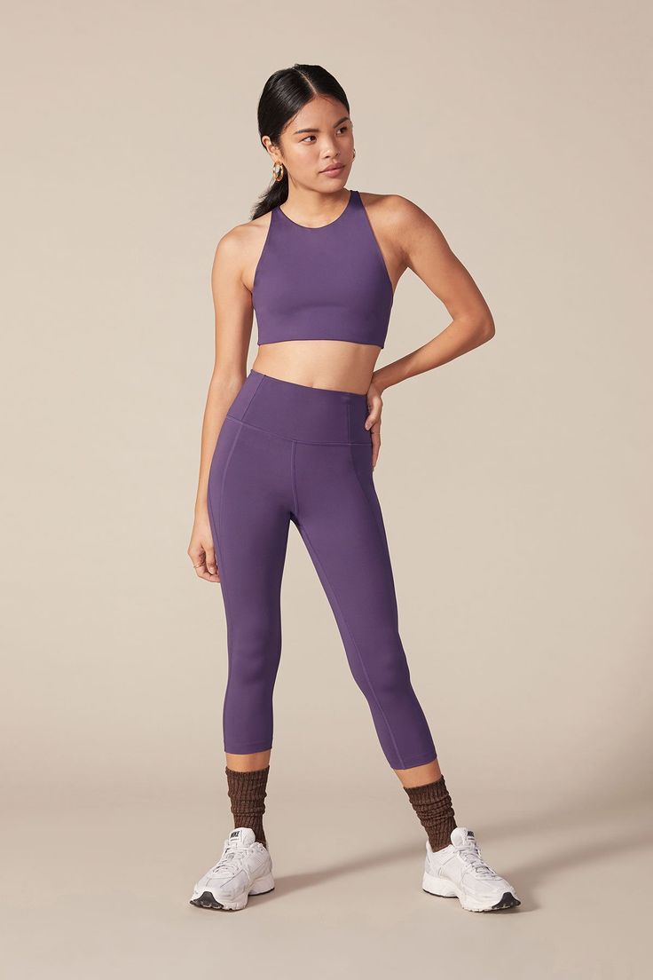 a woman is wearing a purple high waisted leggings and matching sports bra. she is also wearing white sneakers. she is standing with her right hand on her hip.  the background is a light beige color.  she is looking to her right.  she is wearing a casual, athletic outfit perfect for a workout or a run.