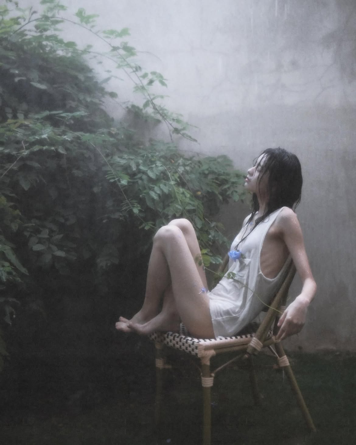 a woman sits barefoot on a wicker chair in a garden during a rain shower. she is wearing a white tank top and looking upwards with a pensive expression. a blue flower is positioned on her top. the background includes lush green foliage and a concrete wall obscured by the rain. the scene has a melancholic and serene atmosphere.