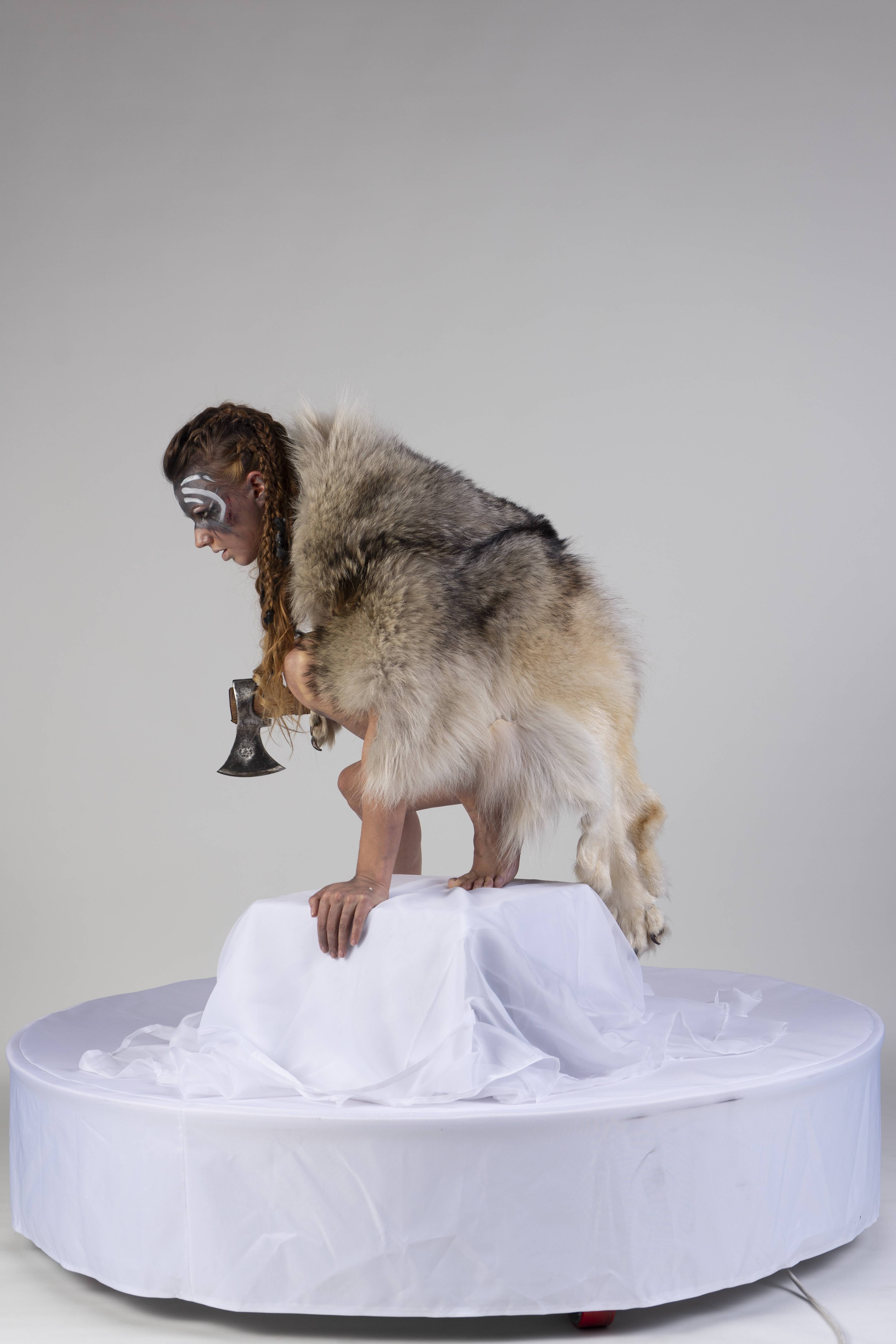 a woman in a fur costume, with face paint and braids, is crouching on a white platform while holding an axe. the scene is set against a plain, light gray backdrop. the costume appears to be made from animal skin or a fur pelt. the platform is covered in white fabric, giving it a stagelike appearance.
