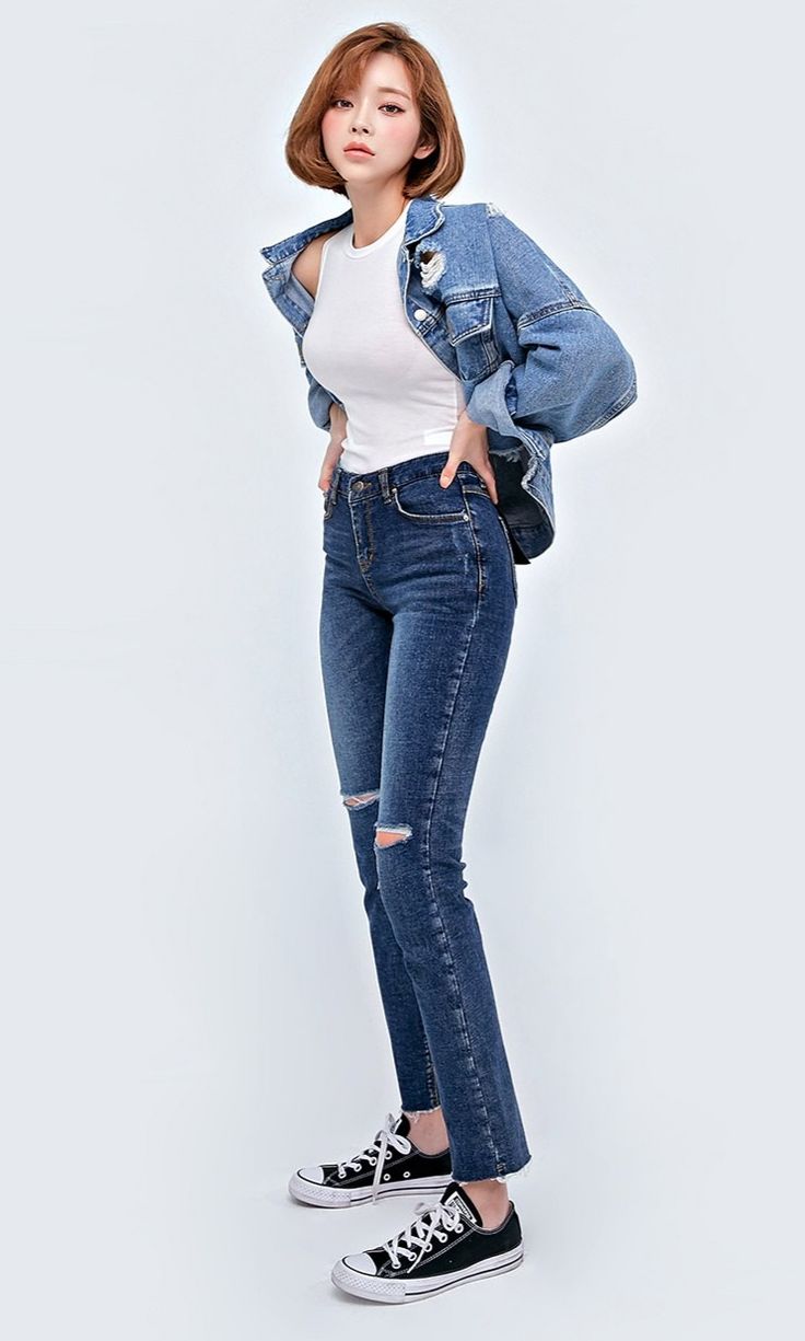a fullbody shot of a young asian woman posing against a white background. she's wearing a white tank top, a distressed light wash denim jacket, dark wash skinny jeans with distressed detailing at the knees, and black converse chuck taylor all star sneakers. her hair is styled in a short bob and she's looking directly at the camera. the overall style is casual and trendy.