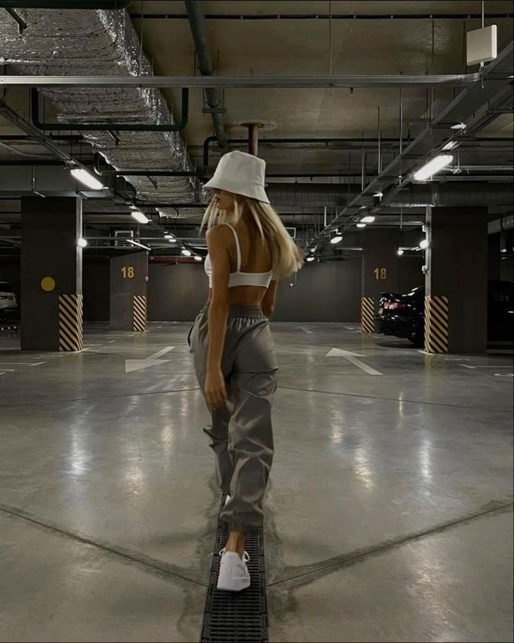 the image shows a young woman from behind, walking down the center of a dimly lit parking garage. she's wearing a white bucket hat, a white crop top, and grey cargo pants. her footwear is white sneakers. the overall aesthetic is dark, moody, and urban. the image is a fashion photograph, showcasing the woman's outfit and style.  the concrete floor and the lines on the garage floor are clearly visible, providing contrast to her outfit.