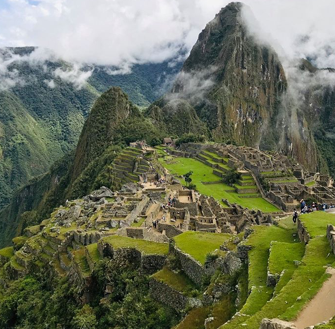 this image captures the ancient city of machu picchu nestled high in the andes mountains of peru. the stone ruins are surrounded by lush green terraces and towering peaks partially obscured by clouds. the scene showcases the historical site's stunning architecture, intricate stonework, and the breathtaking natural landscape. people can be seen exploring the ruins, highlighting its popularity as a tourist destination.