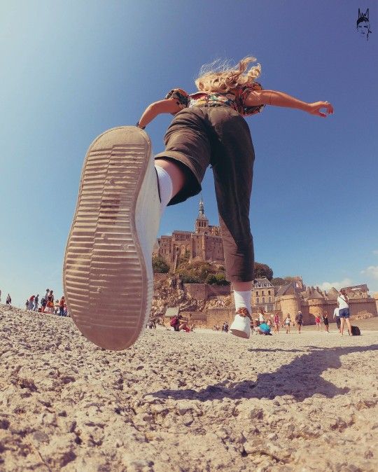 a lowangle perspective shot of a woman jumping with mont saintmichel in the background. the woman has blonde hair and is wearing pants, socks, and shoes. her one leg is raised in midair, showing the sole of her shoe prominently. in the background, the mont saintmichel abbey sits atop a rocky hill. there are also other tourists and buildings visible. the sky is blue. the ground appears to be sandy or rocky.