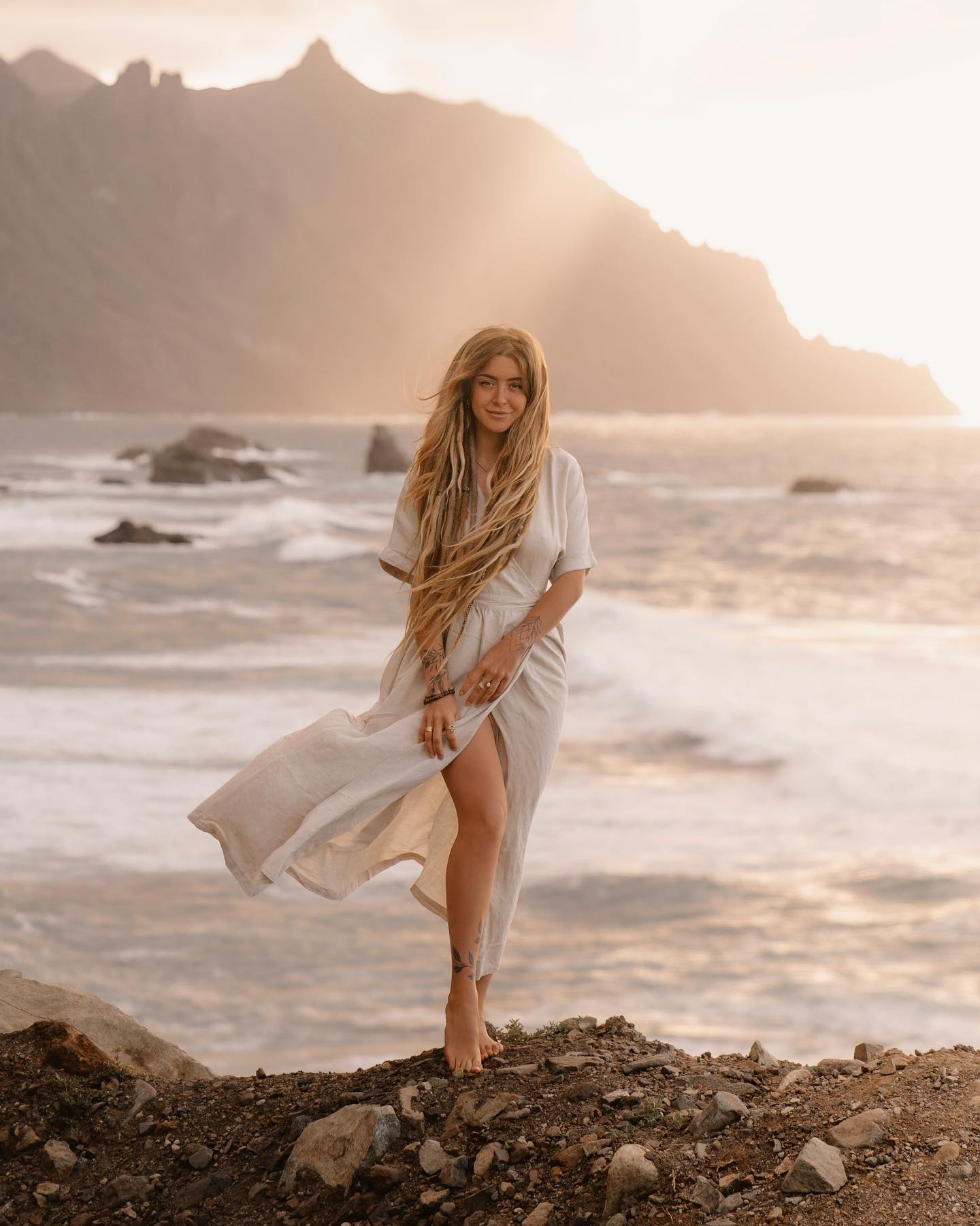 a woman with long blonde dreadlocks is standing on a rocky cliff overlooking a vast ocean. the sunset is casting a warm golden light, illuminating the scene. she is wearing a light beige linen dress that flows in the gentle breeze. the mountains rise in the background, creating a dramatic yet peaceful atmosphere. the overall mood is serene and tranquil, capturing a sense of freedom and connection with nature.