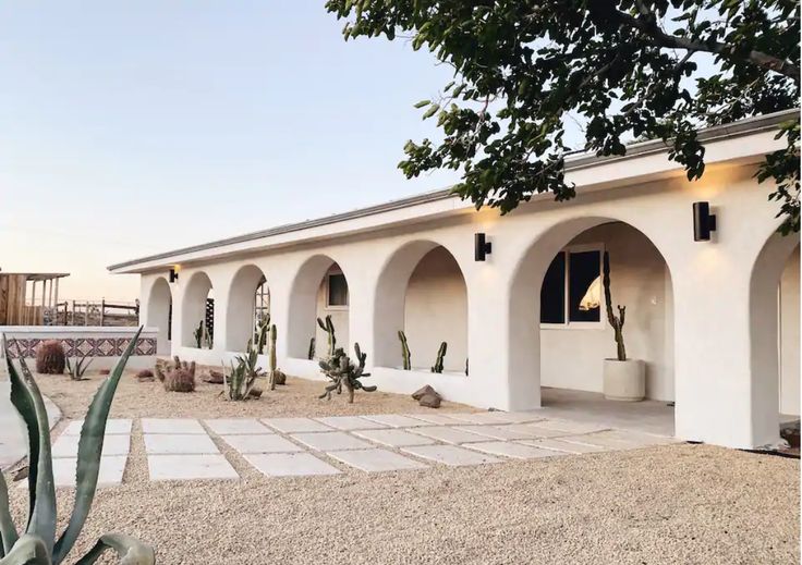 the image showcases a midcentury modern style house nestled in a desert setting.  the architecture features a long, low profile with a series of arched entryways under a simple, flat roof. the exterior is painted white stucco, giving a clean and minimalist aesthetic.  the landscaping complements the architecture, incorporating cacti, succulents, and gravel pathways.  modern, black outdoor sconces are affixed to the exterior walls.  the overall scene projects a feeling of calm and sophistication against a backdrop of desert flora.  a patterned tile section is visible near the entry.