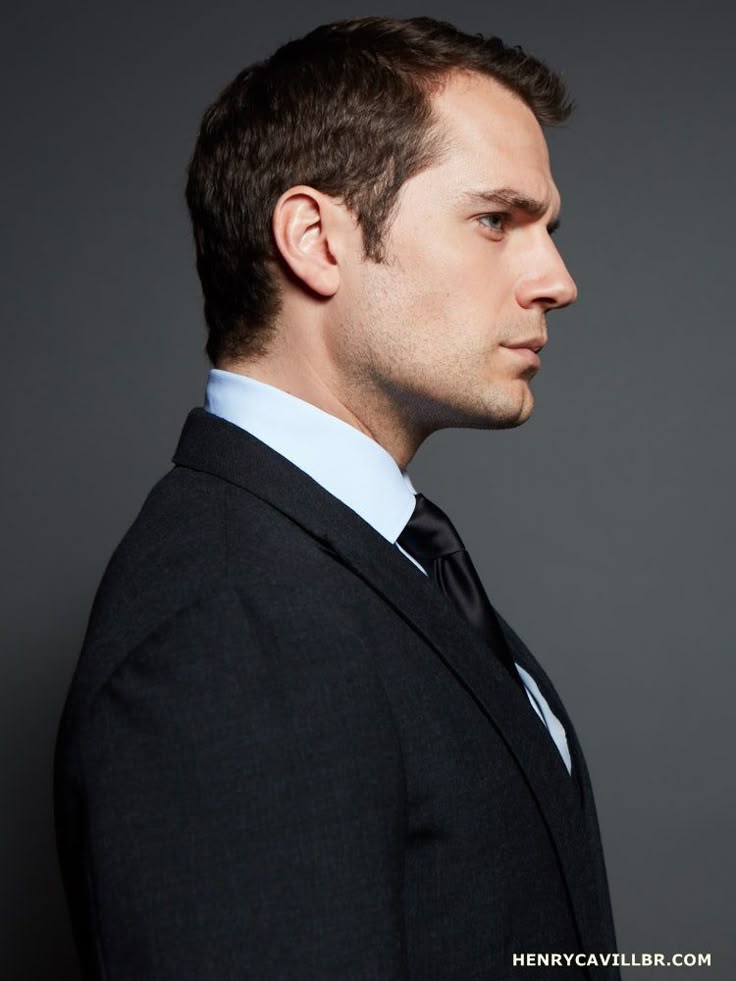 a portrait of henry cavill in a suit and tie, facing to the right. he is wearing a light blue dress shirt underneath a dark gray or black suit. he has short brown hair and a cleanshaven face. the background is a solid gray color. the image is a side view of cavill.
