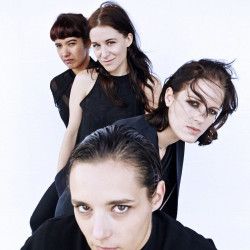 four women, likely a band, pose for a studio portrait. they are dressed in black and white, looking at the camera and smiling.  the photo is set against a white background, and the lighting is soft and diffused.