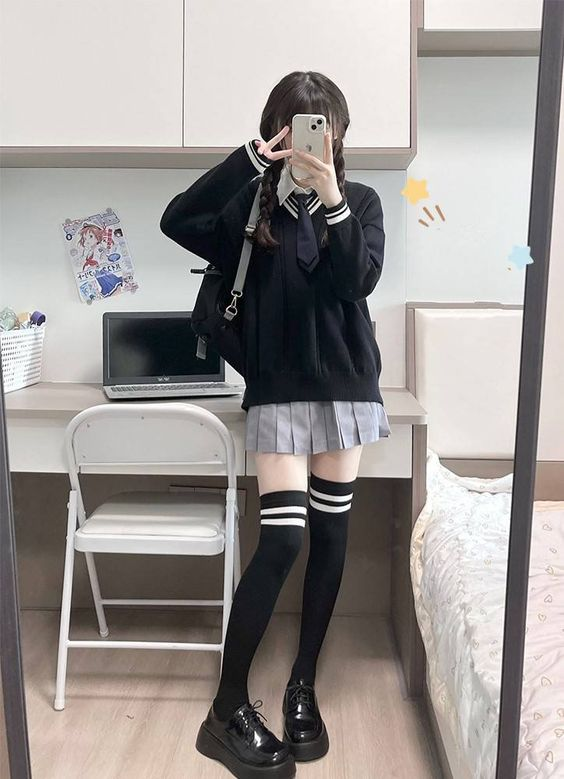 a young woman poses for a mirror selfie in a bedroom. she is wearing a black sweater over a white collared shirt with a black tie, a grey pleated skirt, black and white thigh highs, and black loafers.  the room is simply furnished with a white desk, a white chair, and a bed with grey sheets. the lighting is artificial and creates a bright and clean aesthetic.
