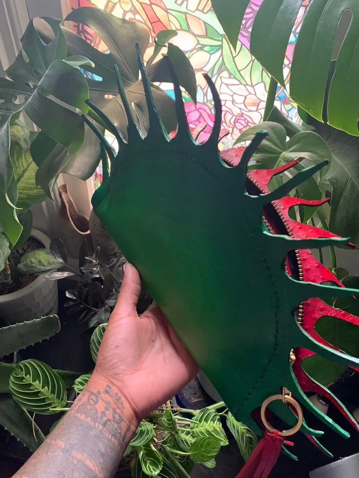 a person is holding a green and red handbag designed to look like a venus flytrap. the handbag is surrounded by various house plants, including a monstera. the person has tattoos on their hand and arm. a stained glass window is visible in the background.
