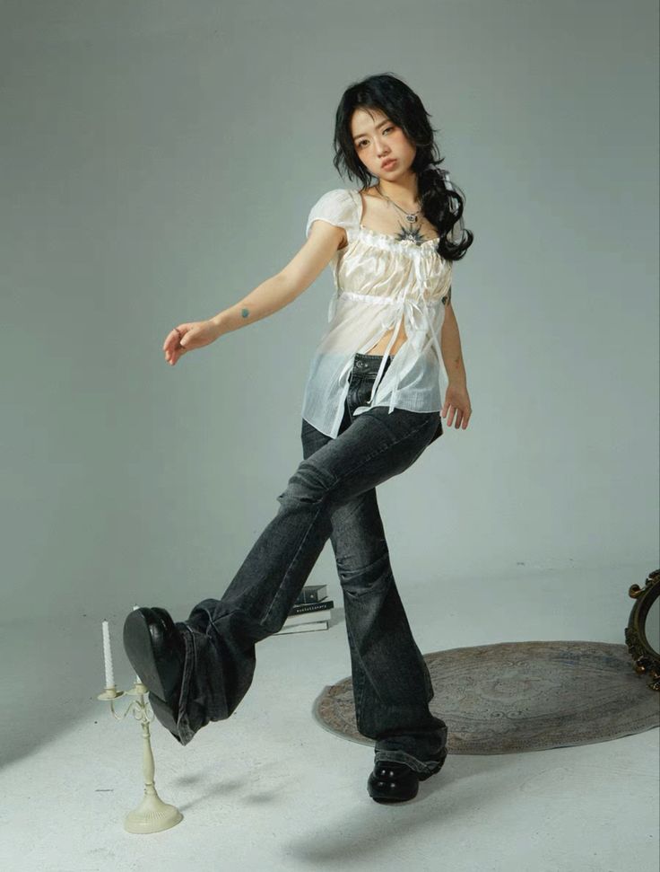 a young woman with long dark hair is posing in a studio against a light gray background. she's wearing a white sheer top with a square neckline and a pair of darkwash bellbottom jeans and black shoes.  her pose is slightly playful and casual; one leg is kicked out. she has a necklace and a visible tattoo on her arm.