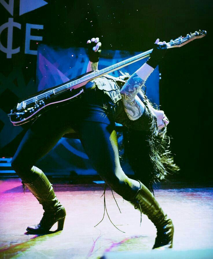 a woman in all black clothing, with long dark hair and tattoos, is playing a bass guitar on a stage during a concert. she is wearing black boots and has a black strap on her wrist. the stage is dark and the lights are shining on her.