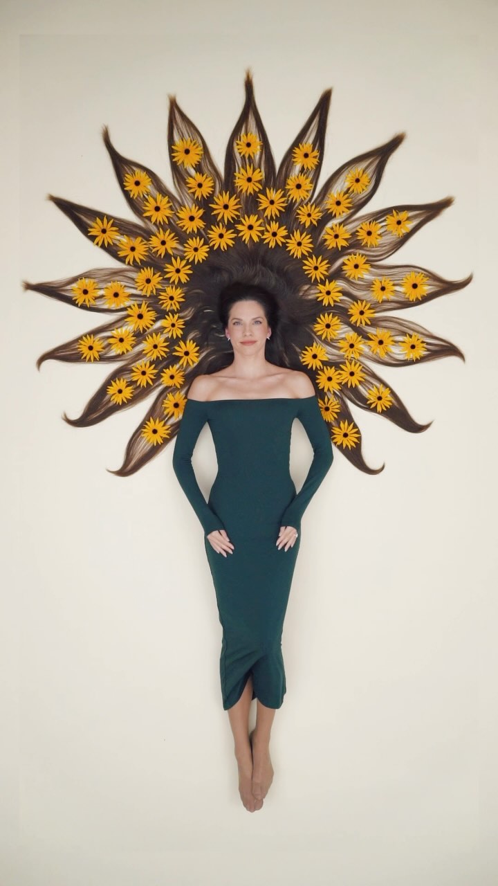 an overhead shot of a woman lying on her back, her long brown hair arranged in a sunburst pattern with blackeyed susans placed between each strand. she is wearing a dark green offtheshoulder dress. the background is a plain light beige. the image is artistic and creative, showcasing both the beauty of the woman and the floral arrangement.
