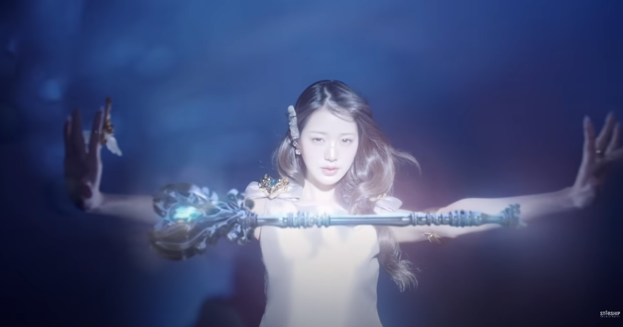 a young woman with long brown hair, wearing a white dress, stands with her arms outstretched holding a glowing staff in front of a blue backdrop. she appears to be in a performance setting, possibly for a music video or concept photo, and is styled with an ethereal and powerful aesthetic.