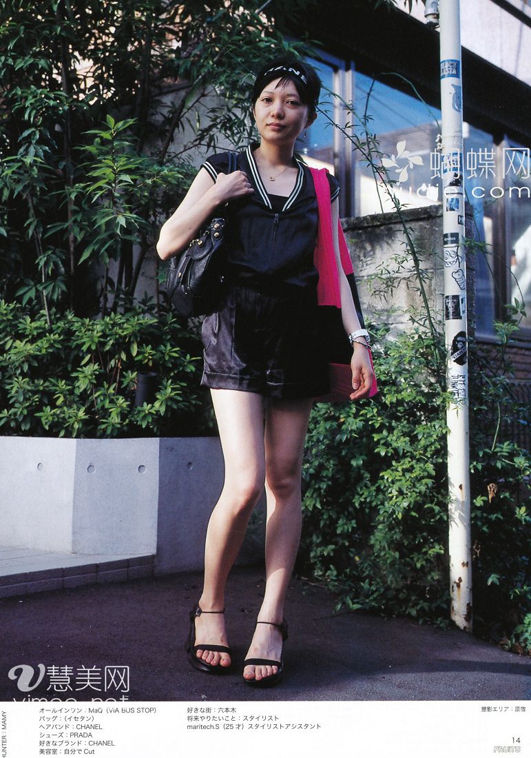 a young japanese woman is posing for a fashion magazine on a city street. she is wearing a black romper with a pink top underneath, black strappy sandals, and a black headband. she has a black handbag slung over her shoulder. she is standing on a sidewalk in front of some greenery and a pole with japanese writing on it. the photo is welllit with natural daylight.