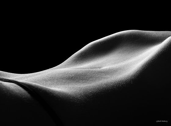 the image is a black and white closeup photograph of a woman's body. the lighting is dramatic, using lowkey lighting to highlight the curves and textures of the skin.  the composition is abstract, focusing on the flowing lines and forms of the body rather than explicit detail.  the overall mood is sensual and artistic, emphasizing the beauty of the human form through light and shadow. the background is completely black, drawing all attention to the subject. the photo appears to be a fine art print with a photographer's signature visible.