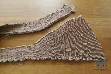 two pieces of pleated fabric are lying on a wooden surface. the fabric appears to be beige or light brown, and the pleats are tightly packed. one piece is a long strip, while the other is a wider, fanshaped piece. a logo labeled "larva" is on the bottom right corner.