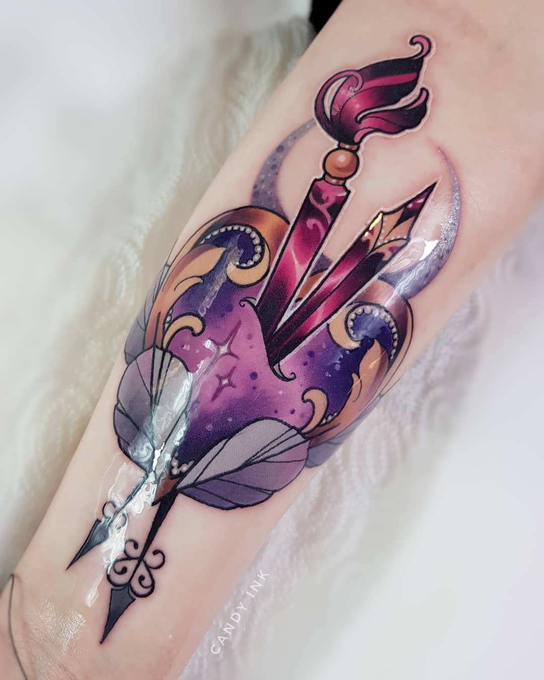 a tattoo of two paint brushes crossed over each other to create the shape of an arrow piercing through a heart. the heart has a cosmic and galaxy theme with purples, pinks, and blues. there are gold sparkles on the heart as well.  the feathers of the arrow are below the heart.  the tattoo is on someone's forearm.