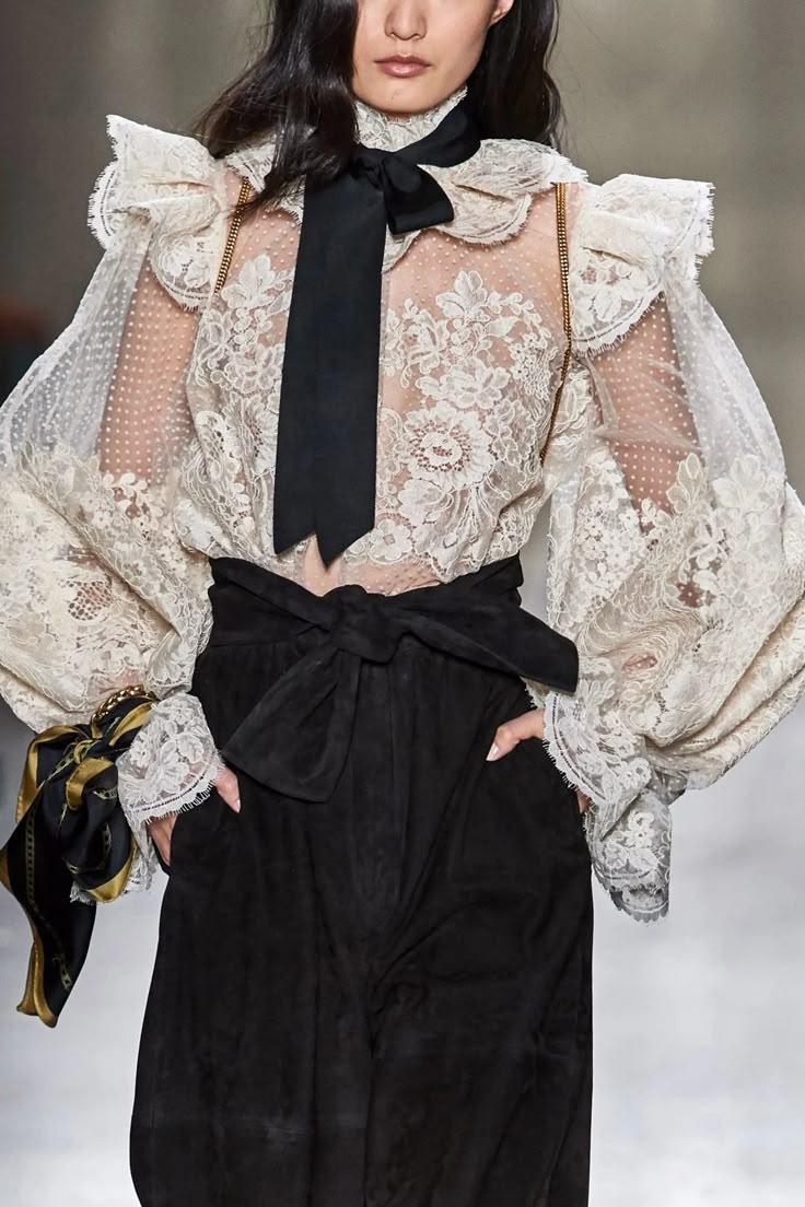 a woman is shown from the chest down wearing a lace blouse with a black bow around her neck and a pair of highwaisted black pants. the blouse is creamcolored with intricate lace detailing and features puffy sleeves with ruffled edges. a black bow tie is loosely tied around the neck, contrasting against the lace. the black pants cinch at the waist with a wide black sash tied into a bow. the woman has her hands casually placed in her pockets. the background appears to be a simple studio setting. the overall aesthetic is romantic and vintageinspired.