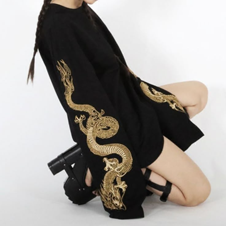 a woman is kneeling wearing a black oversized long sleeved shirt with gold embroidered dragon designs down the sleeves. she is wearing platform shoes and a black garter on her thigh. her hair is in braids.