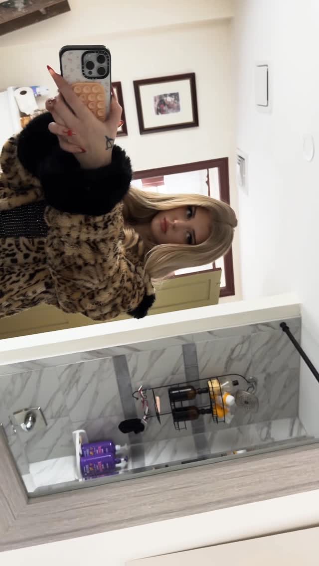 a woman takes a selfie in the mirror in a bathroom. she is wearing a leopard print coat. the phone in her hand has a tan phone case. behind her are picture frames on the wall, the shower, shampoo, and bottles.