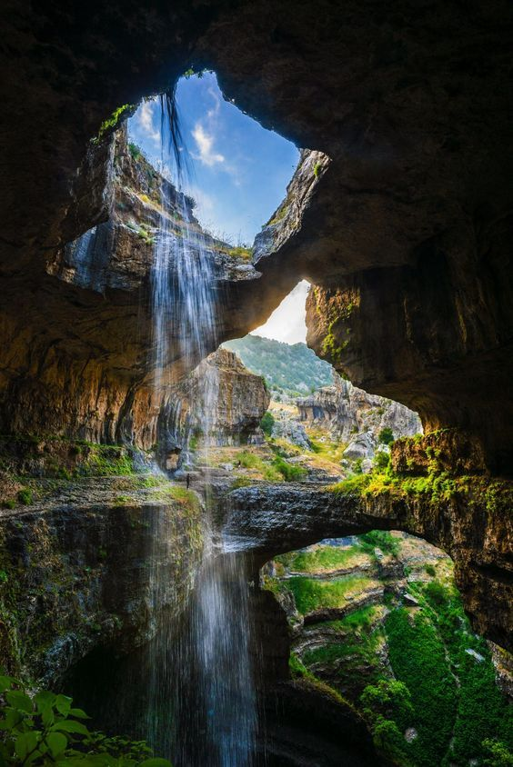 the image showcases the baatara gorge waterfall in tannourine, lebanon, as seen from within a cave. a powerful waterfall plunges through a sinkhole in the cave's ceiling, creating a stunning visual spectacle. the water cascades over rocky cliffs and formations, eventually flowing into a deep gorge. lush greenery surrounds the area, enhancing the natural beauty of the landscape. the sky is visible through the opening in the cave's ceiling, allowing sunlight to illuminate the scene.