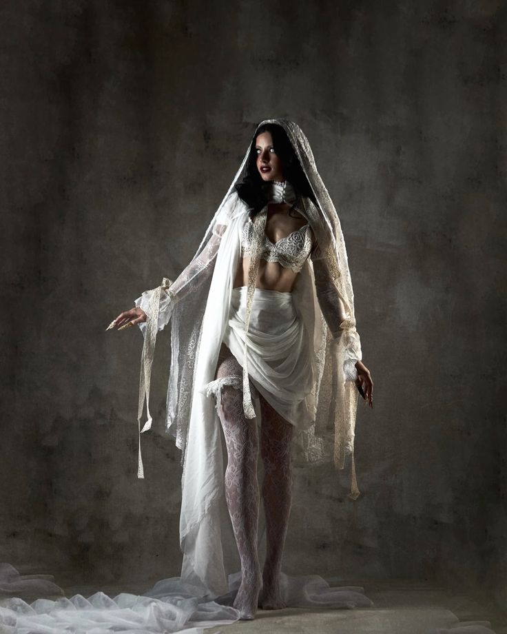 the image is a dramatic, fulllength portrait of a woman dressed in a gothicinspired bridal attire. she wears a flowing white veil and gown with intricate lace detailing, which contrasts with a visible lace lingerie set underneath.  the dress is loosely draped, revealing parts of her legs in lace stockings. her dark hair and makeup enhance the overall dark and mysterious atmosphere. the background is a textured, dark grey wall, providing a stark contrast to the white of her clothing. the lighting is dramatic, focusing on the model and creating a moody, almost ethereal ambiance. the overall style evokes a sense of gothic romance or a dark fairytale.