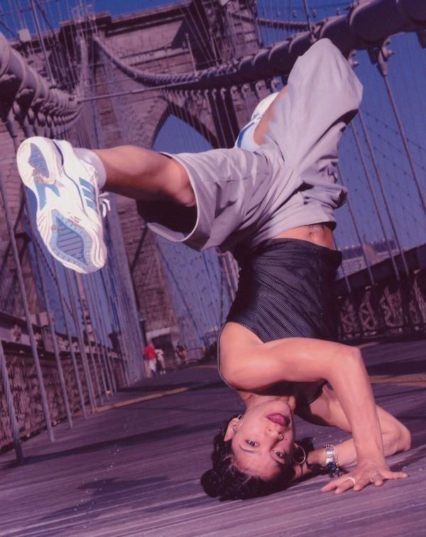 a young woman wearing a black shirt, gray shorts, and white sneakers, is upside down while breakdancing on a bridge. she is smiling and looks confident.