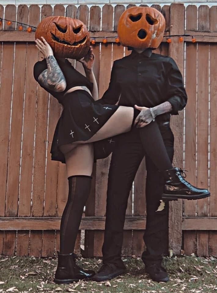 a couple dressed in spooky halloween costumes. the female is wearing a black skirt with crosses and a pumpkin head. the male is wearing all black and has a pumpkin head on as well.  they're posing in front of a wooden fence, with the man holding the woman's leg up. they are both smiling and enjoying their costumes.