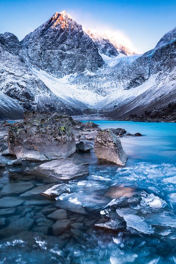 a scenic shot of a snowcovered mountain range reflecting into a vibrant blue lake with scattered rocks and patches of ice.